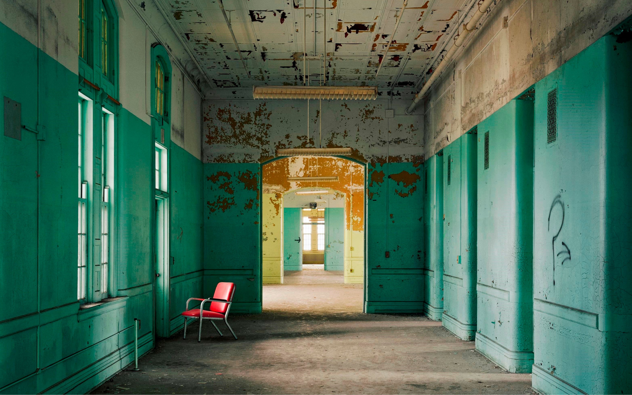 Derelict institutional hallway with peeling green paint and lone red chair in front of window.